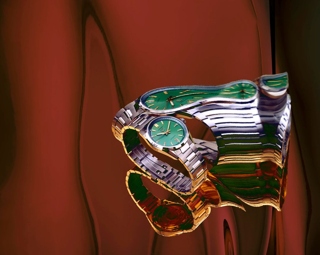 Close-up of a wristwatch with a metal strap and green face distorted by a reflective surface on a maroon background. Timepiece.
