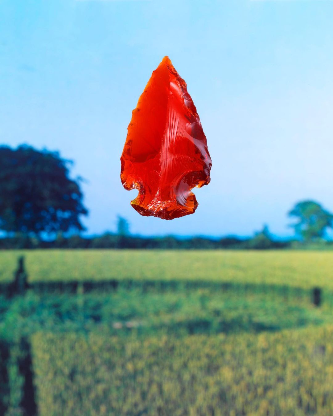 Reddish-orange arrowhead artifact centered over a blurred green field and pale blue sky background in a studio shot.