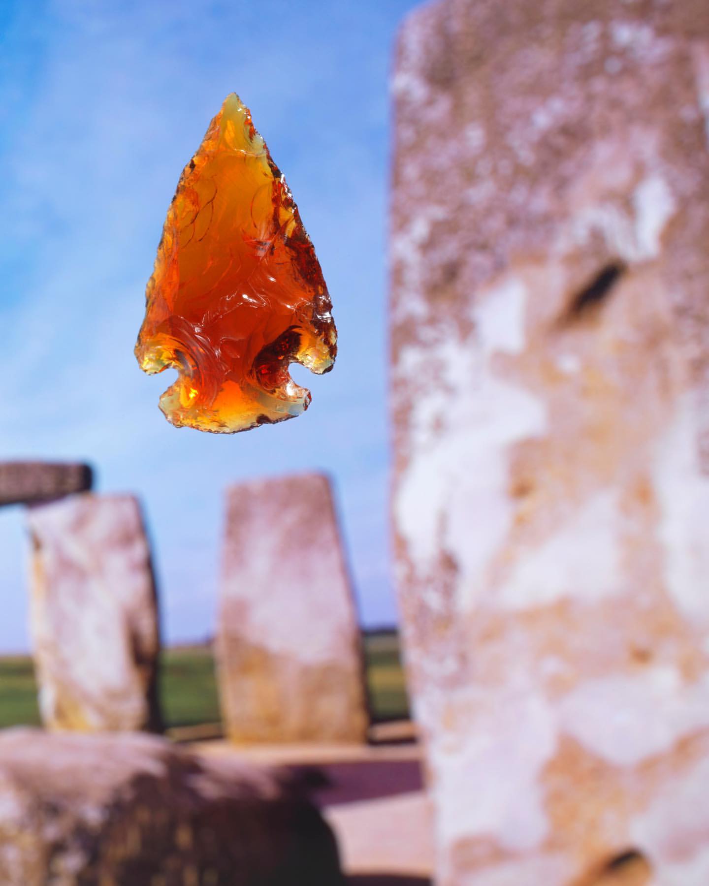 Amber-colored arrowhead made of obsidian stone, points upward in front of weathered stone columns against a light blue sky.