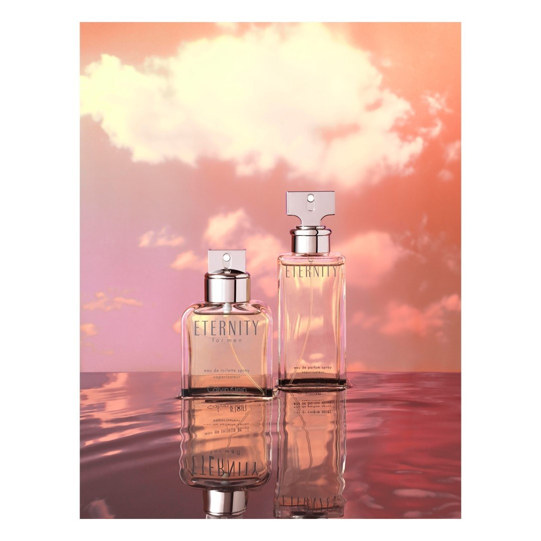 Two Calvin Klein Eternity for Men cologne bottles are reflected in rippled water against a sunset cloudscape backdrop.