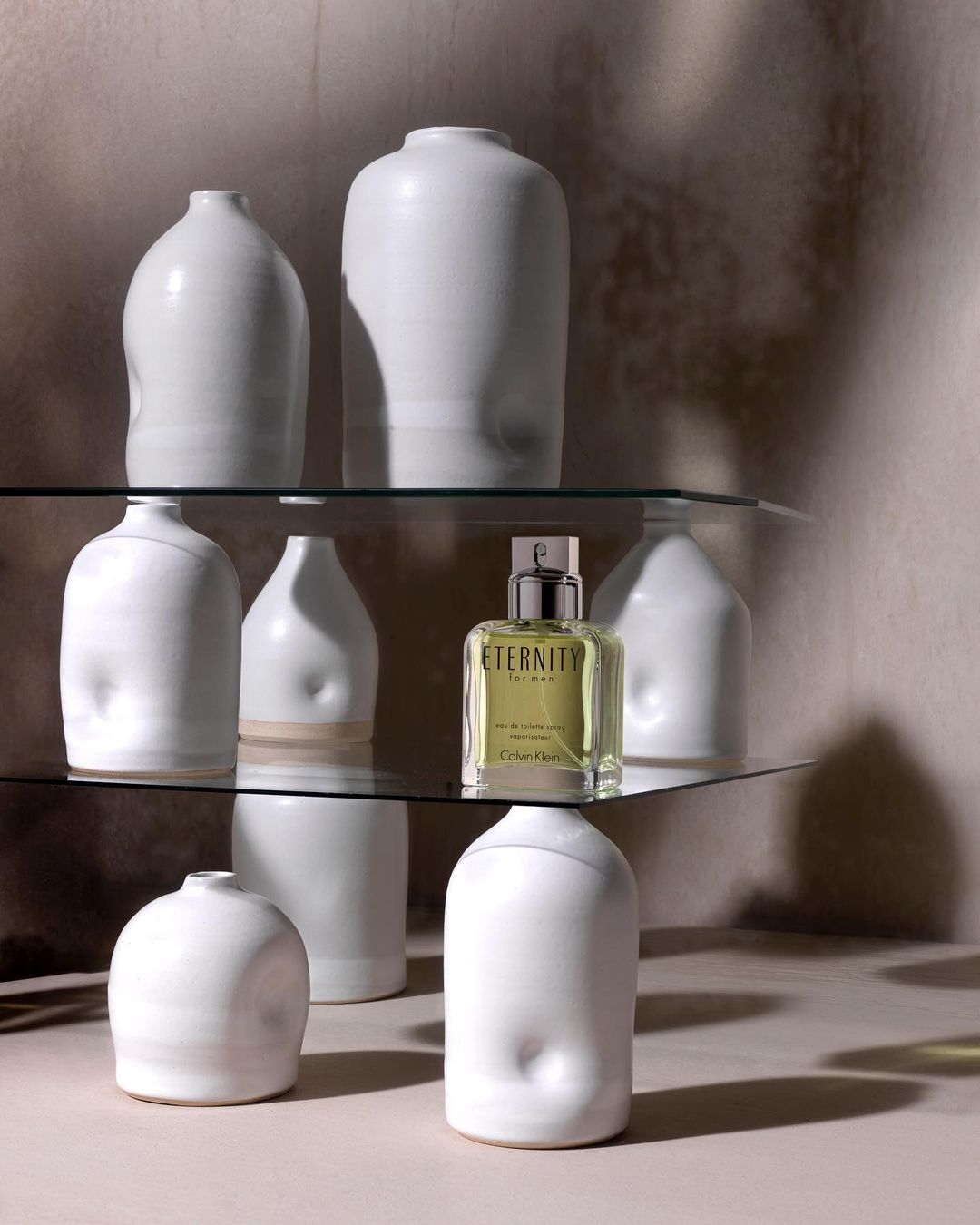 Perfume bottle of Eternity by Calvin Klein on a glass shelf surrounded by modern white vases in front of a neutral colored wall.