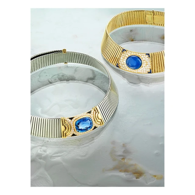 Two gold and silver ribbed choker necklaces with blue gemstones displayed on a white marbled surface in a studio setting.