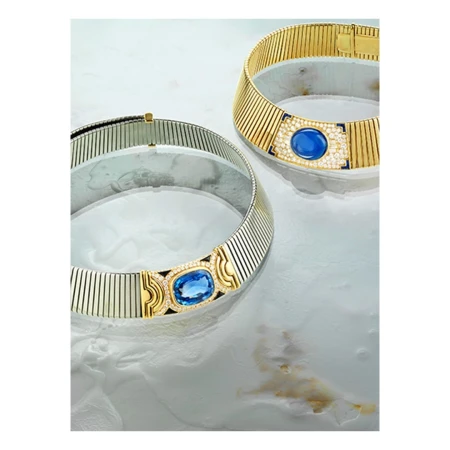 Two gold and silver ribbed choker necklaces with blue gemstones displayed on a white marbled surface in a studio setting.