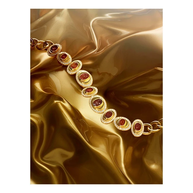 Close-up of a golden chain necklace with oval pendants featuring red stones and diamond accents, set against a draped gold fabric backdrop.