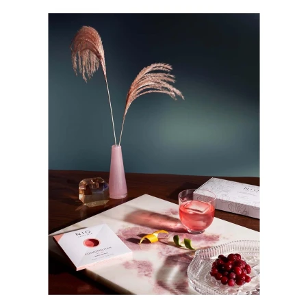 Drink photography featuring a Cosmopolitan cocktail, vase with pampas grass, and garnishes on a marble surface with dark backdrop.