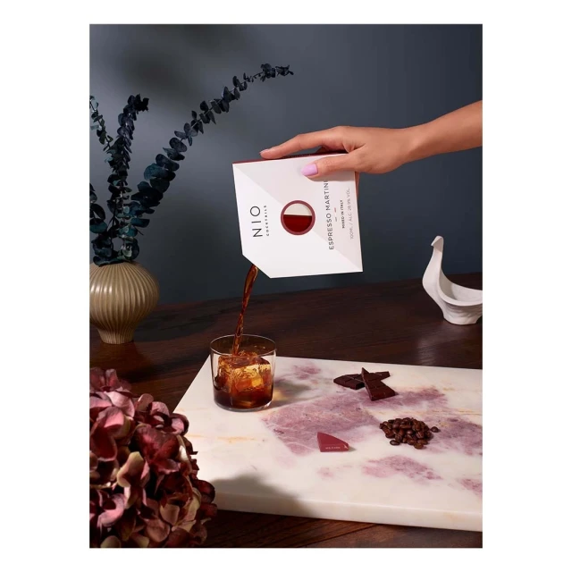 Espresso martini cocktail being poured into a glass with ice from a white container on a marble serving board, next to coffee beans and chocolate.
