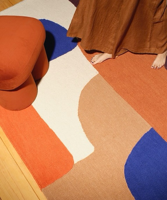 Eye-level indoor shot of a woman in a dress, a colorful abstract rug, and a rust-colored stool with tripod legs on wood flooring.