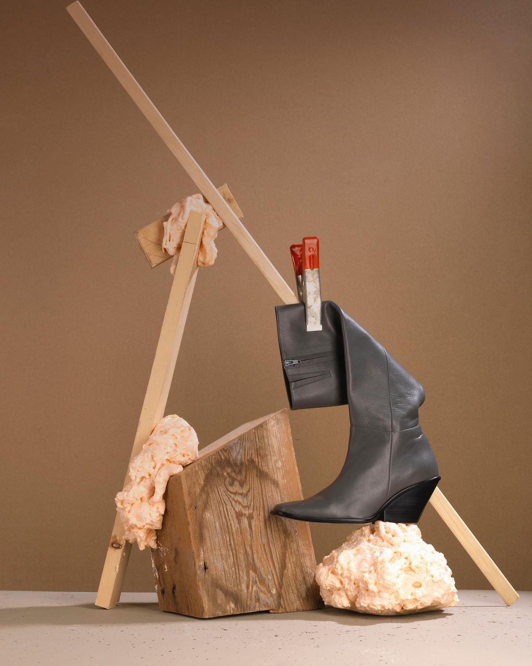 Studio shot of a gray leather boot suspended by red clothespins, balanced on wooden block and foam against a beige backdrop.