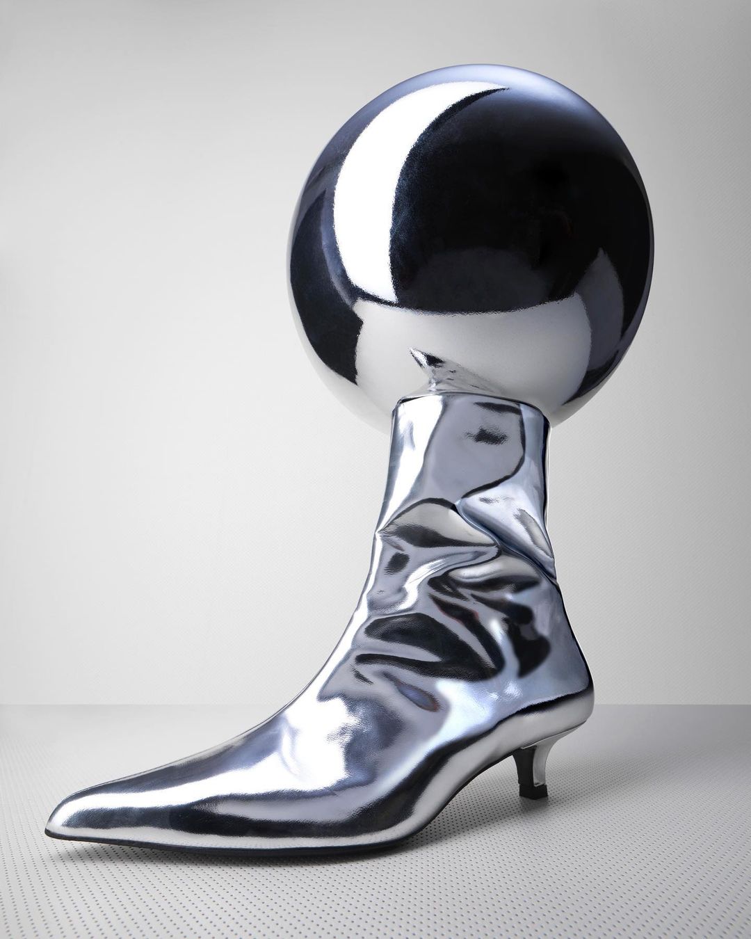 Still life shot of a shiny silver ankle boot under a reflective silver globe on a seamless white background in a studio setting.