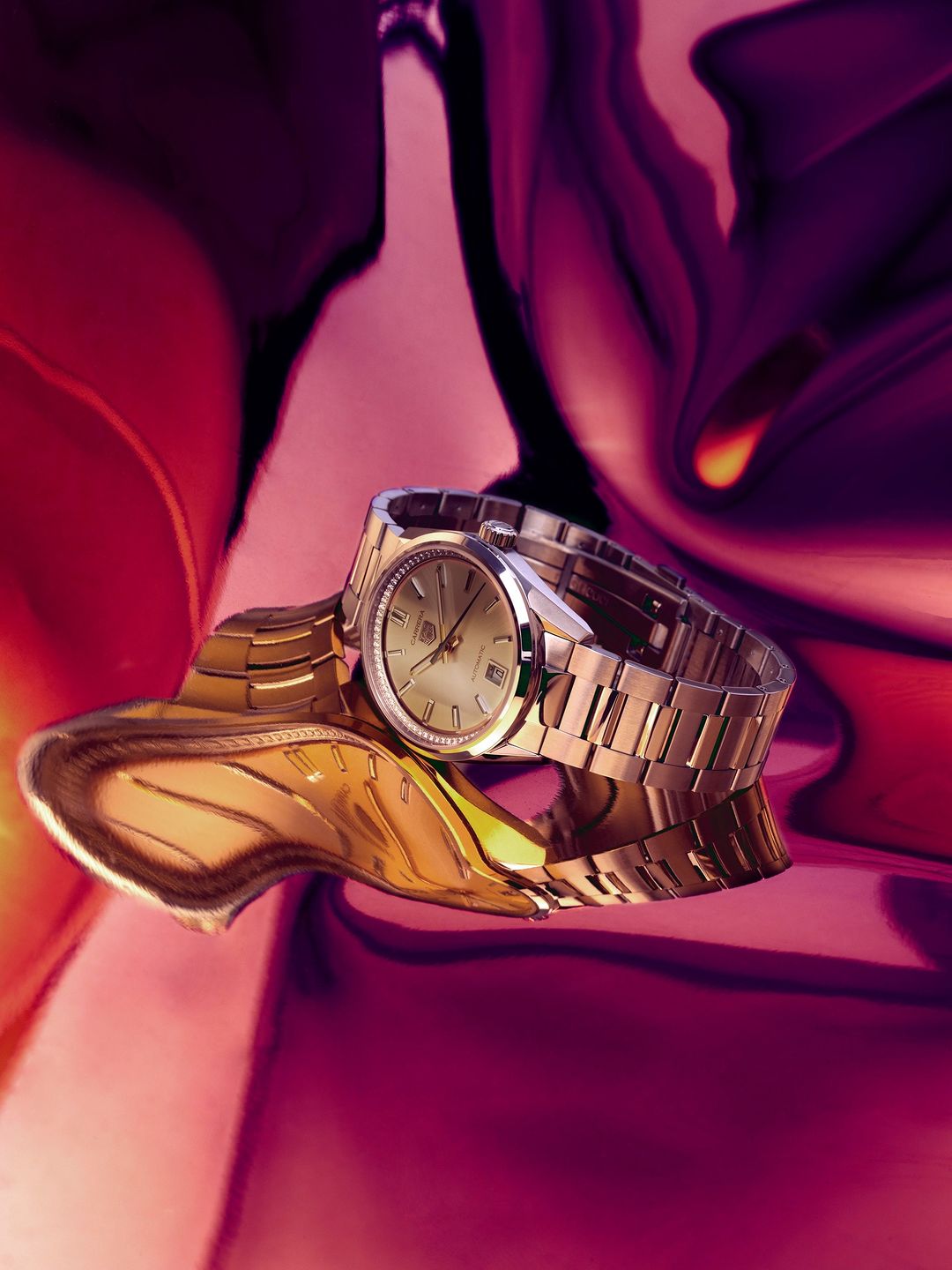 Elegant gold wristwatch on a textured gold metallic surface with a blurred swirling pink and purple background in a studio setting.