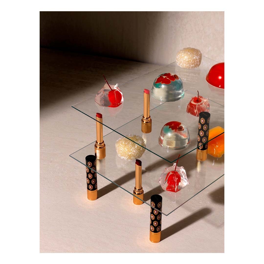 Elegant beauty still life: lipsticks with golden accents arranged on glass shelves alongside jello and gummy candies in a studio setting.