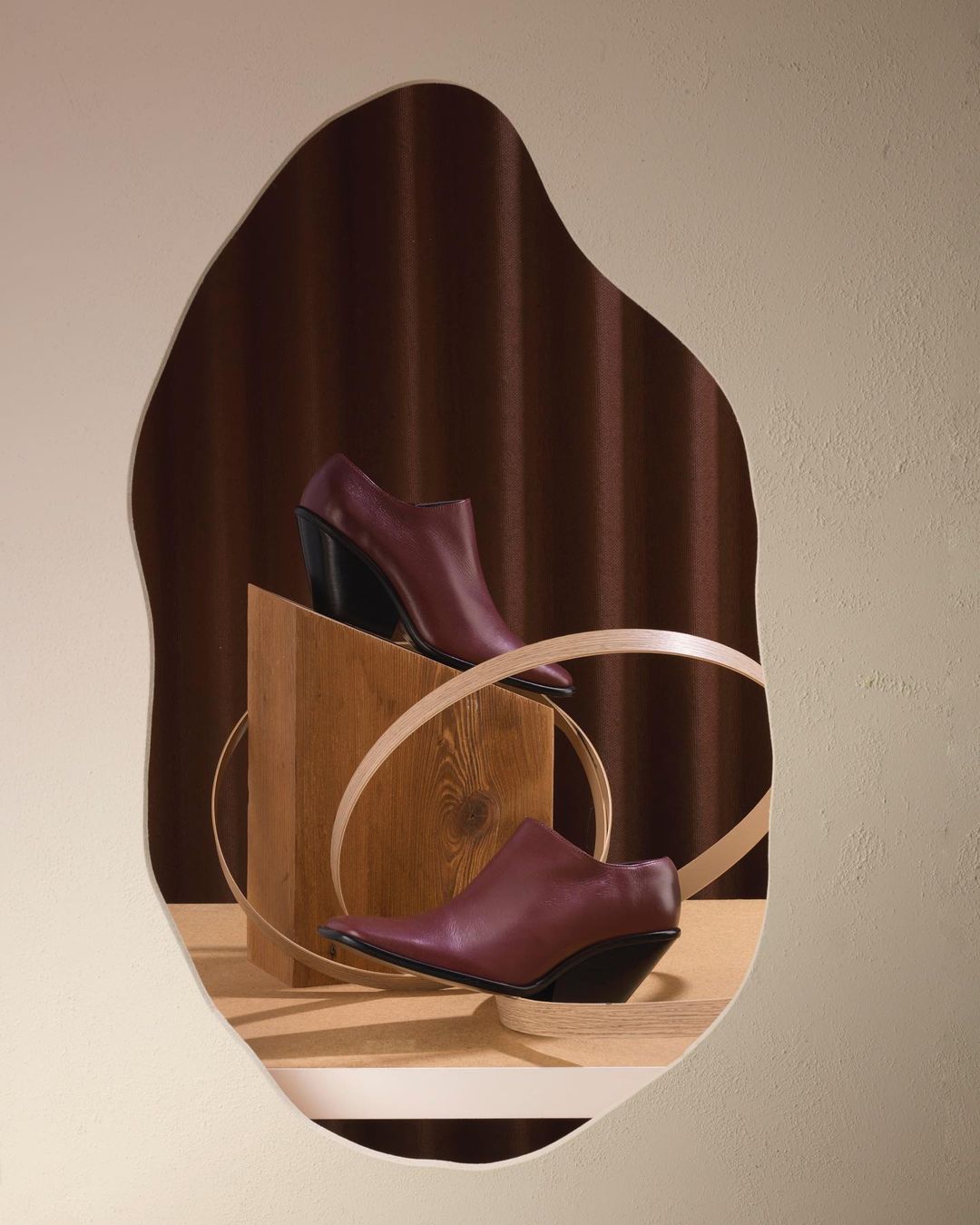 Elevated still life featuring burgundy ankle boots with black heels, displayed on wooden blocks and surrounded by neutral ribbons.