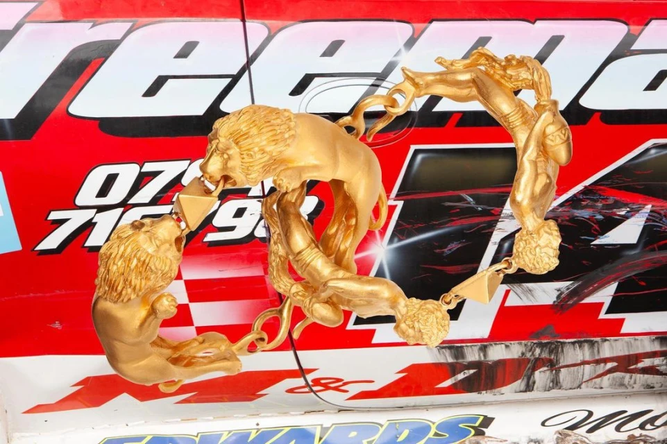 Golden sculpture featuring lions and human figures affixed to the side of a race car with a red, white, and black color scheme.