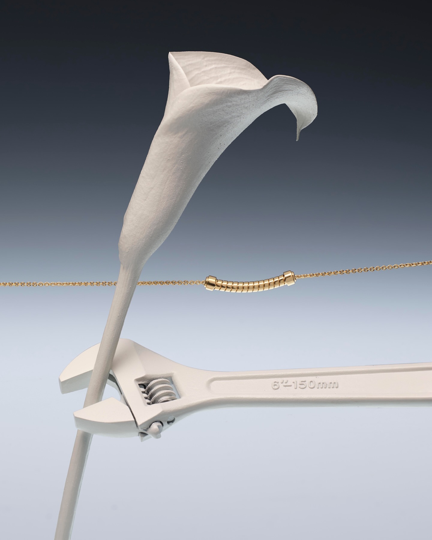 Still life of a white calla lily being held by a white wrench, with a gold necklace and dark gray to light gray background gradient.