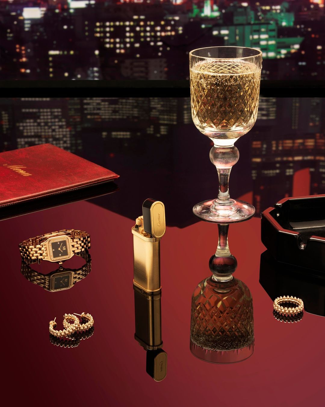 Elegant still life featuring a glass of champagne, gold jewelry, lighter, and a red notebook against a blurred city nightscape.