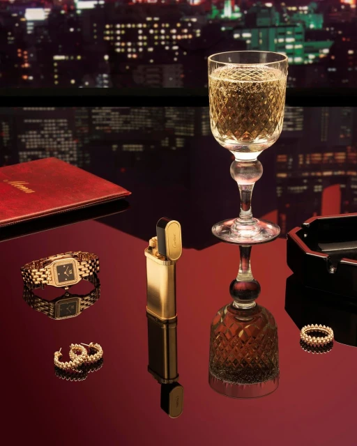 Elegant still life featuring a glass of champagne, gold jewelry, lighter, and a red notebook against a blurred city nightscape.