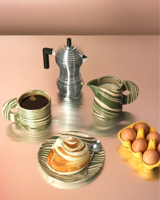 Still life of a coffee pot, coffee cup, pastry, cream pitcher, eggs, and spoon on a metallic surface with a peach backdrop.