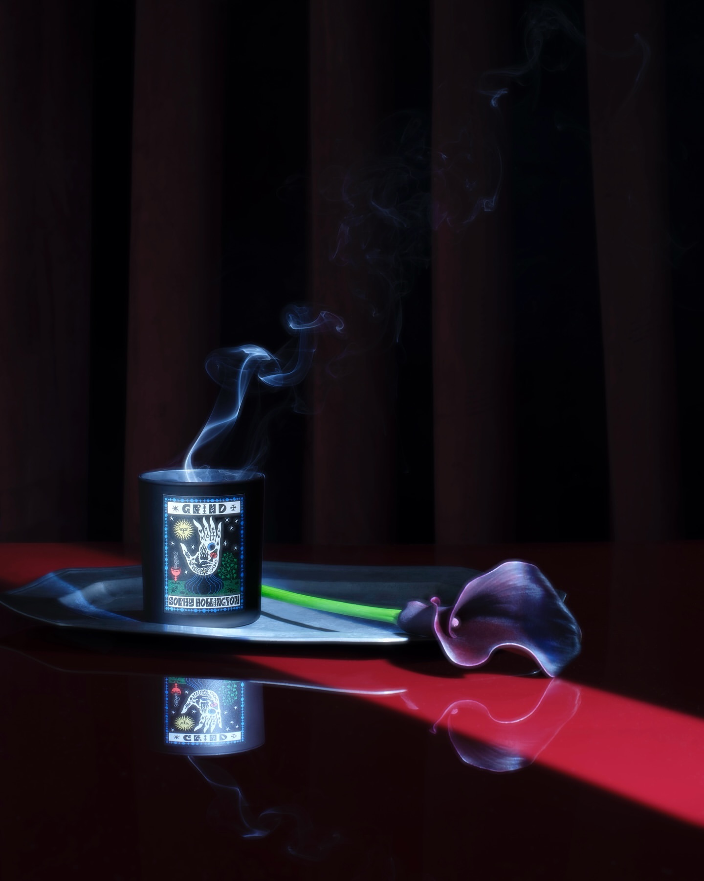 Black candle in a glass jar with a whimsical illustration, wisps of smoke rising, and a dark calla lily on a silver tray.