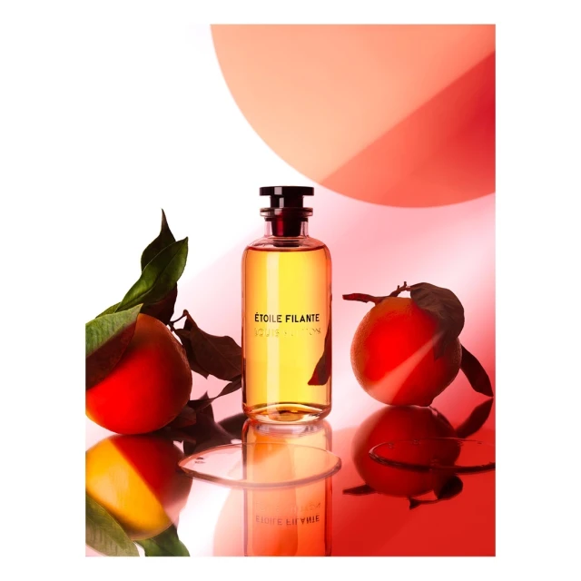 Perfume bottle of Etoile Filante Louis Vuitton on a reflective surface, complemented by two oranges and a coral circle backdrop.