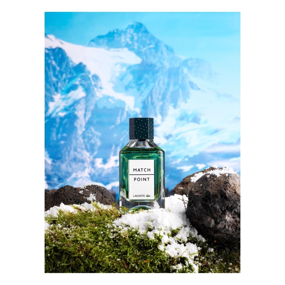 Lacoste Match Point fragrance bottle prominently displayed on mossy terrain with light snow, set against a backdrop of majestic snow-capped mountains.
