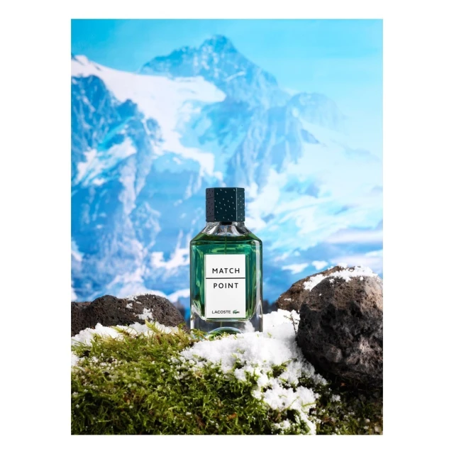Lacoste Match Point fragrance bottle prominently displayed on mossy terrain with light snow, set against a backdrop of majestic snow-capped mountains.