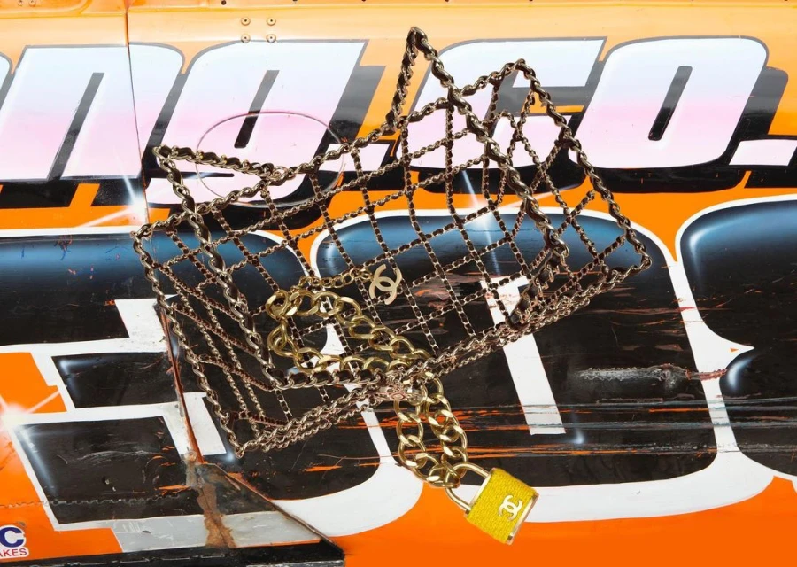 A chain-link handbag with the Chanel logo is displayed on an orange racing vehicle, offering a juxtaposition of luxury and sport.
