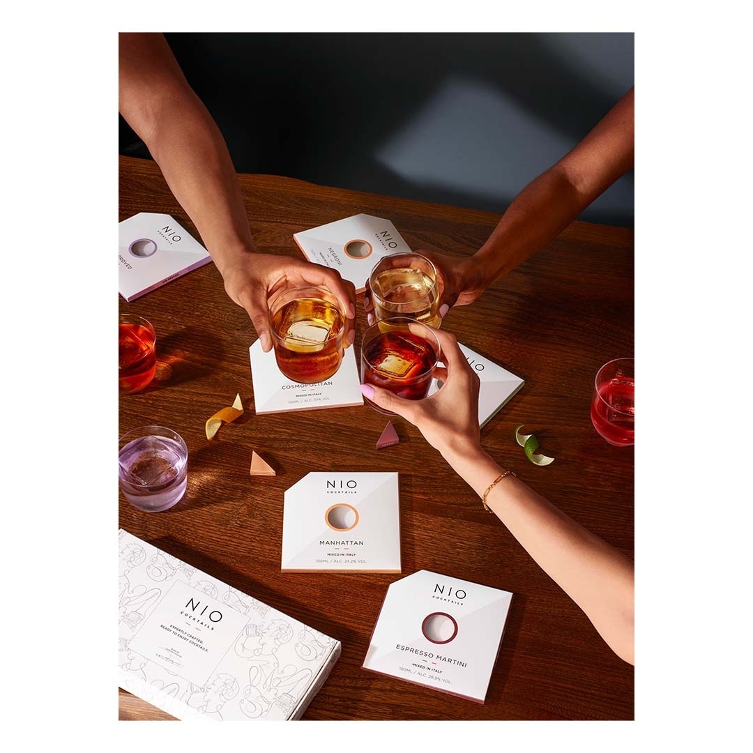 People toasting cocktails with pre-mixed NIO cocktail boxes on a wooden surface. Drinks are garnished with orange and lime peels.