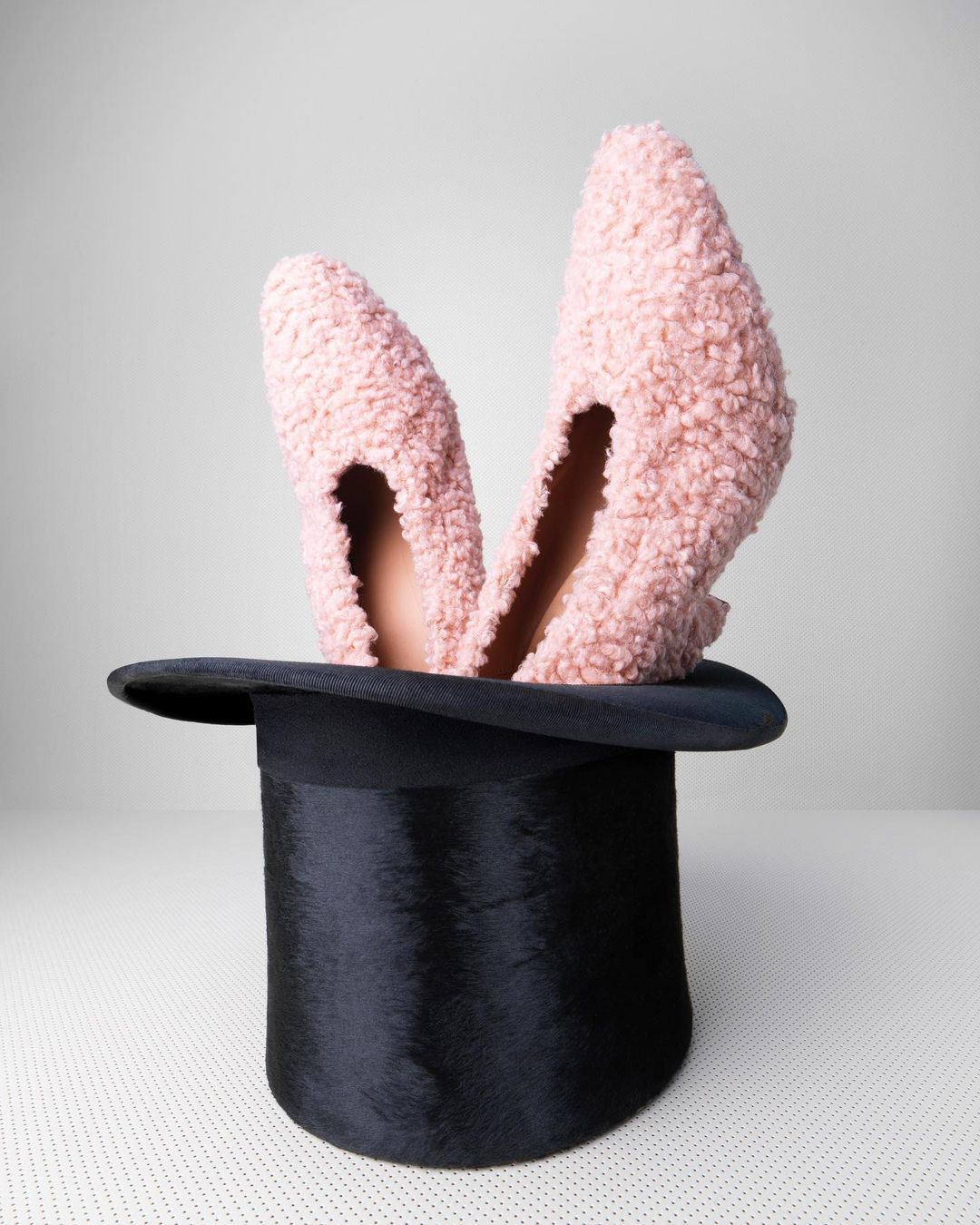 Pink furry high-heeled shoes inside a black top hat on a white surface, creating a whimsical and fashionable composition with studio lighting.