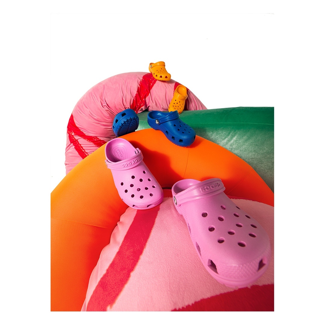 Colorful Crocs footwear in pink, blue, yellow, and orange are creatively arranged atop layered fabric surfaces in a studio setting.