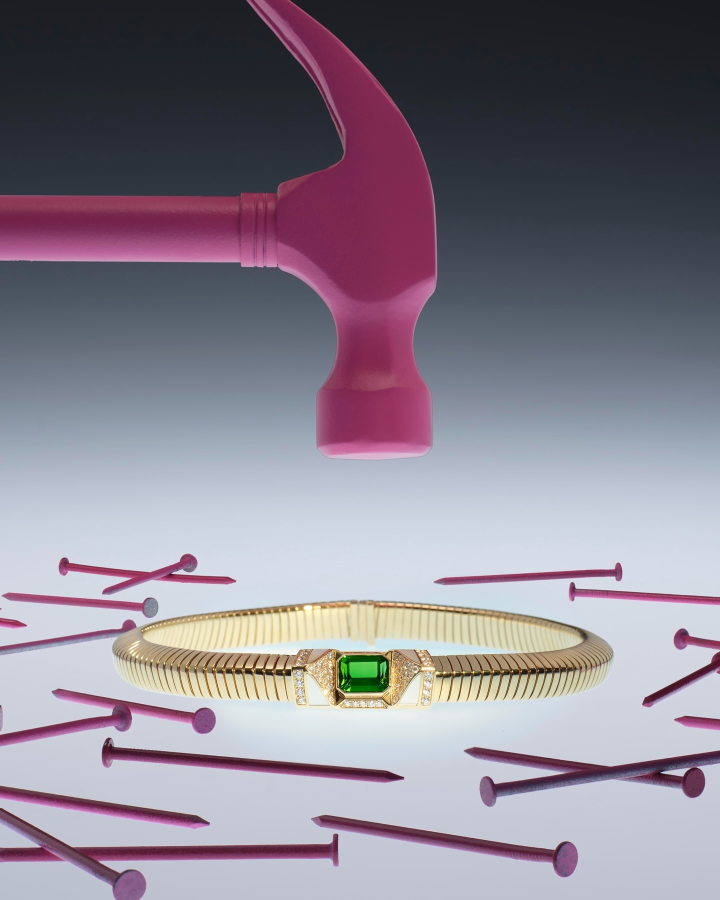 Still life shot of gold bracelet with green gem encircled by pink nails, and pink hammer poised above it against a gray background.