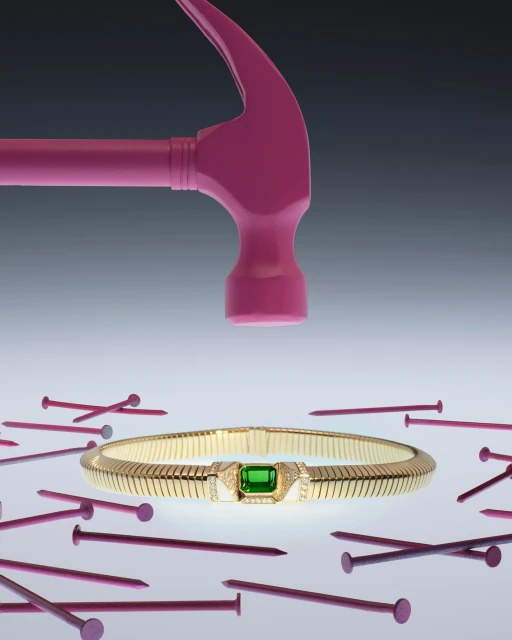 Still life shot of gold bracelet with green gem encircled by pink nails, and pink hammer poised above it against a gray background.