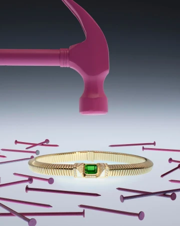 Still life shot of gold bracelet with green gem encircled by pink nails, and pink hammer poised above it against a gray background.