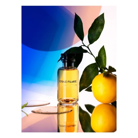 A glass perfume bottle labelled Etoile Filante next to a fresh yellow orange with green leaves on a reflective surface.