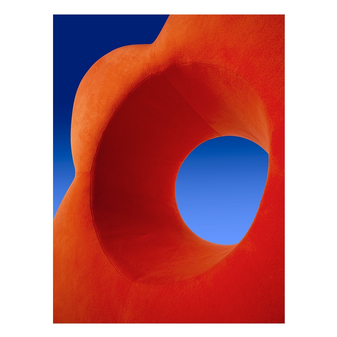 Abstract orange sculpture with a circular opening set against a bright blue sky, creating a strong contrast and a modern aesthetic.
