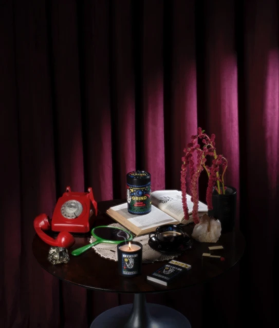 Still life photo on a dark wooden table: a jar, candle, landline phone, book, flowers, ashtray, hand mirror, and cards with a dark maroon curtain backdrop.