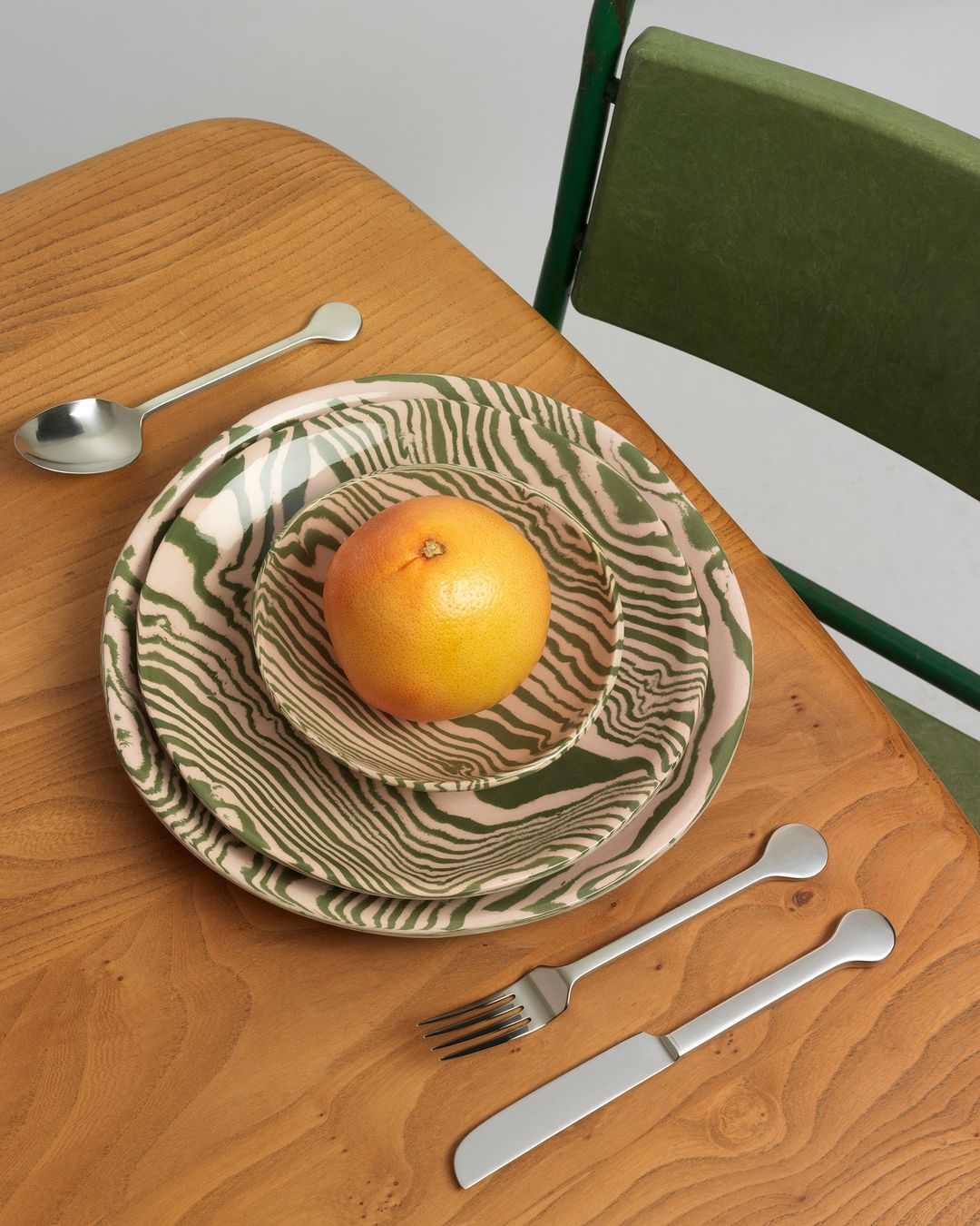 Overhead shot on a wooden table with a grapefruit on patterned plates, a fork, a knife, and a spoon, near an olive green chair.