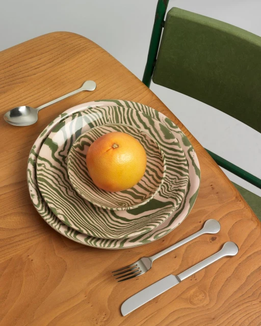 Overhead shot on a wooden table with a grapefruit on patterned plates, a fork, a knife, and a spoon, near an olive green chair.