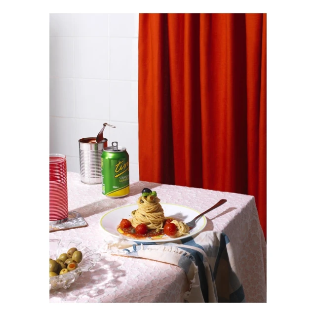Still life photograph of a plate of spaghetti with cherry tomatoes and basil next to a green beverage can, on a table with a white tablecloth.