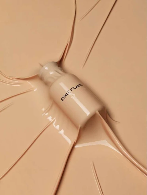 Makeup bottle surrounded by cosmetic foundation. Beige background, beauty product shot. ETOILE FILANTE text on the bottle.