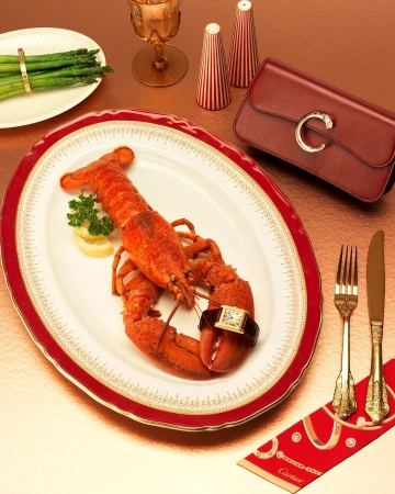 Overhead shot of cooked lobster, asparagus, and luxury accessories on a gold surface with red rimmed plate and Cartier branding.