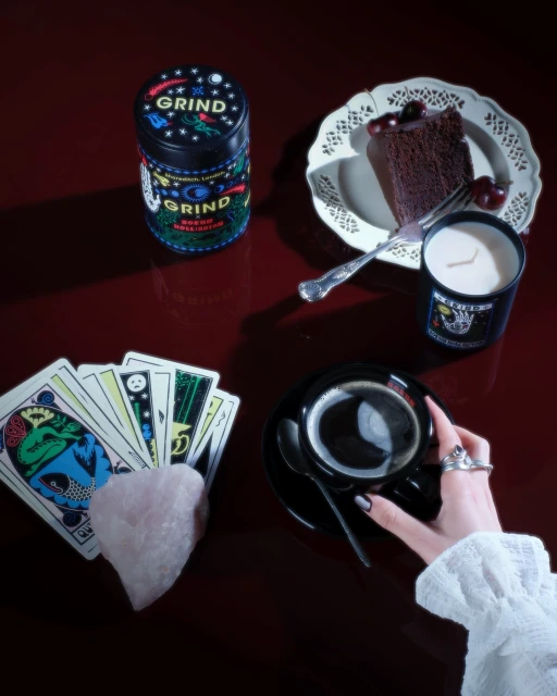Still life featuring tarot cards, a Grind coffee tin, cake, a candle, and a cup of coffee on a dark red surface. 