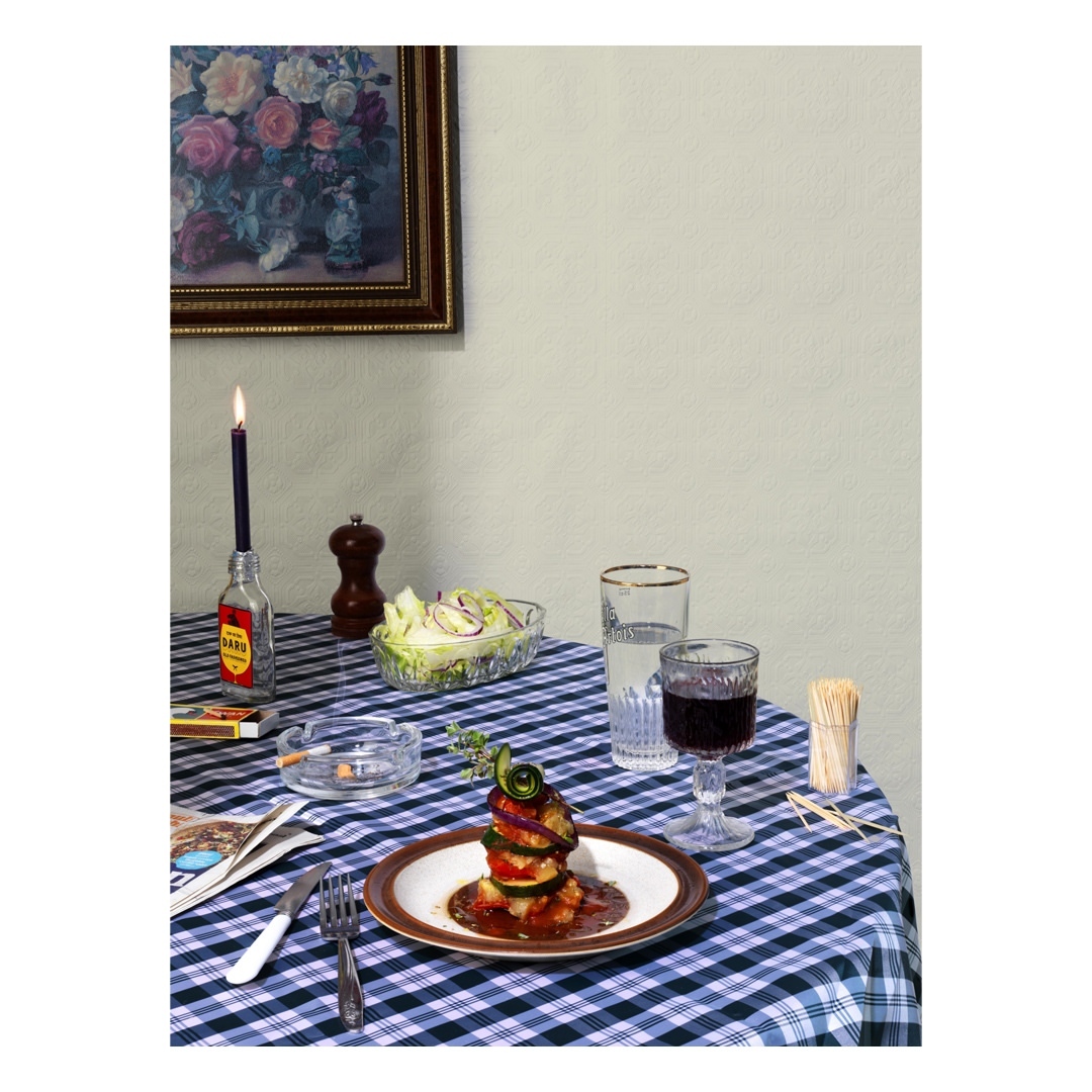 Still life shot of a plated zucchini tower with wine, salad, candle, cigarettes, and matches on a blue and white checkered tablecloth.