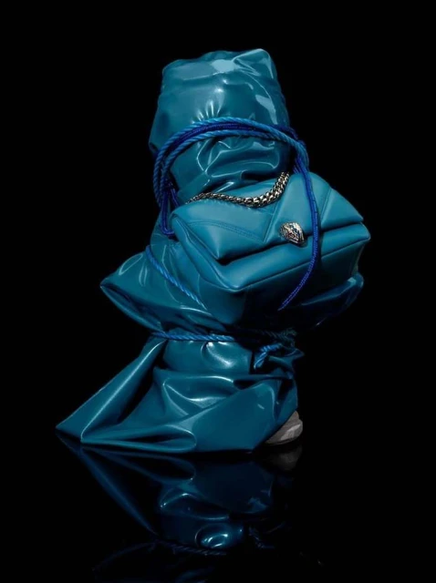 Close-up of a blue Bulgari handbag wrapped in blue plastic and rope, sitting on a reflective black surface in a photo studio.
