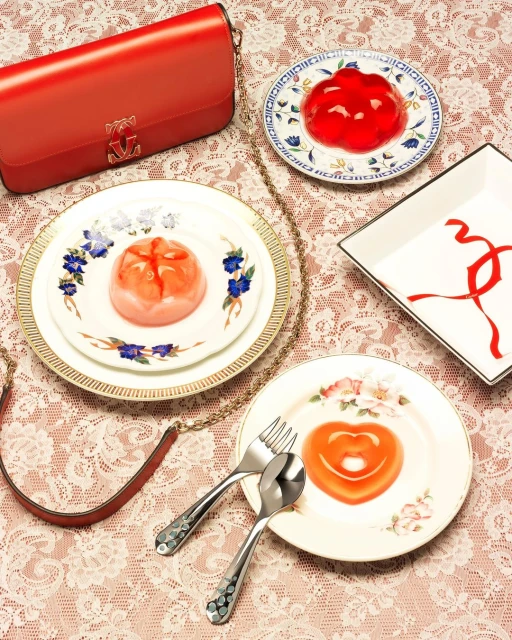 Overhead still life of jellies on plates, with silverware, a red purse with a gold chain strap, and a lace tablecloth.