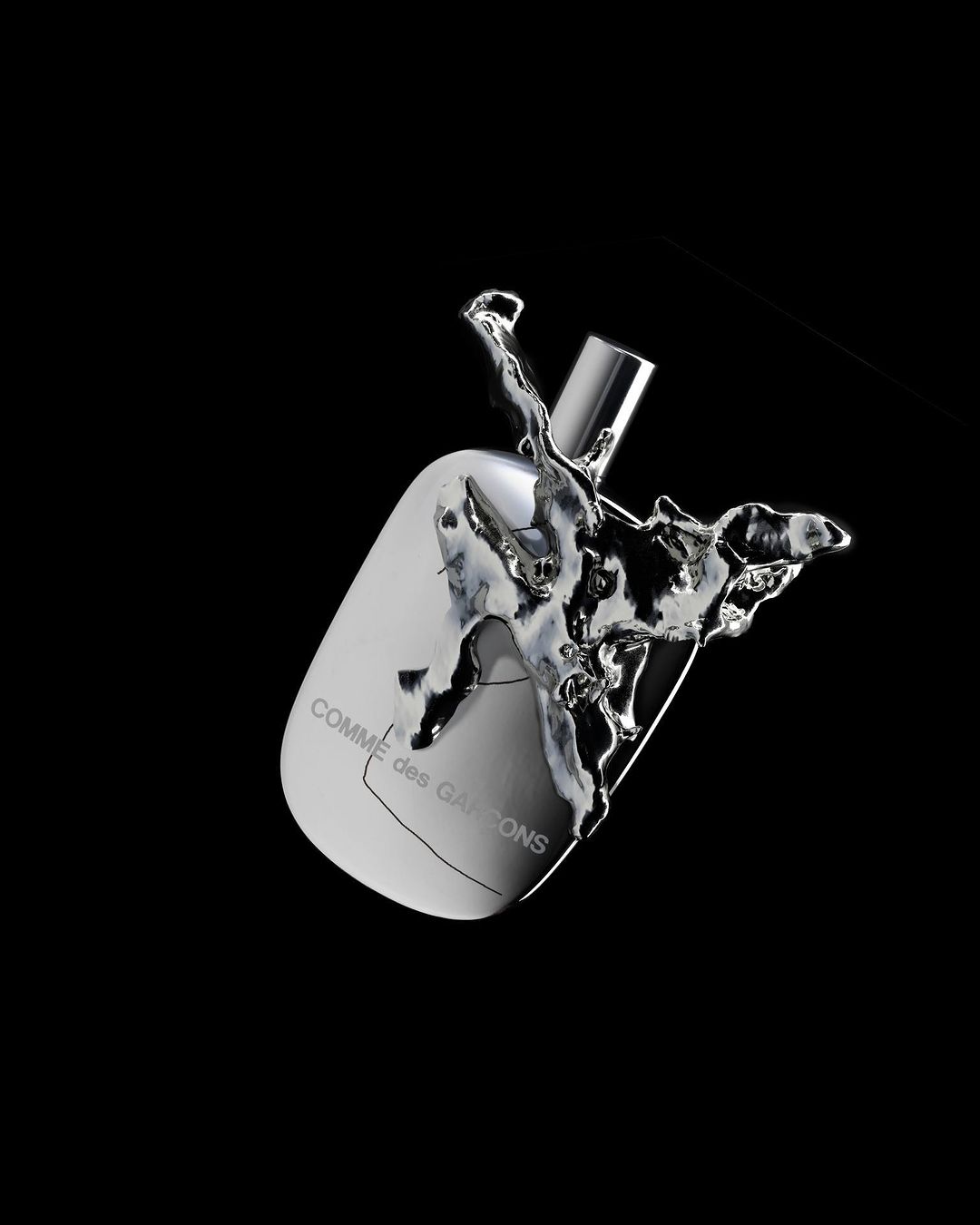 Studio shot of a reflective silver perfume bottle with liquid splashing around it on a black background, displaying the COMME des GARÇONS logo.