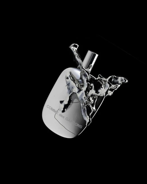 Studio shot of a reflective silver perfume bottle with liquid splashing around it on a black background, displaying the COMME des GARÇONS logo.