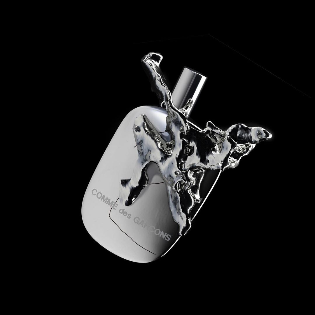 Comme des Garçons perfume bottle angled, surrounded by splash of silver liquid against a stark black background in a studio setting.