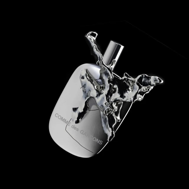 Comme des Garçons perfume bottle angled, surrounded by splash of silver liquid against a stark black background in a studio setting.