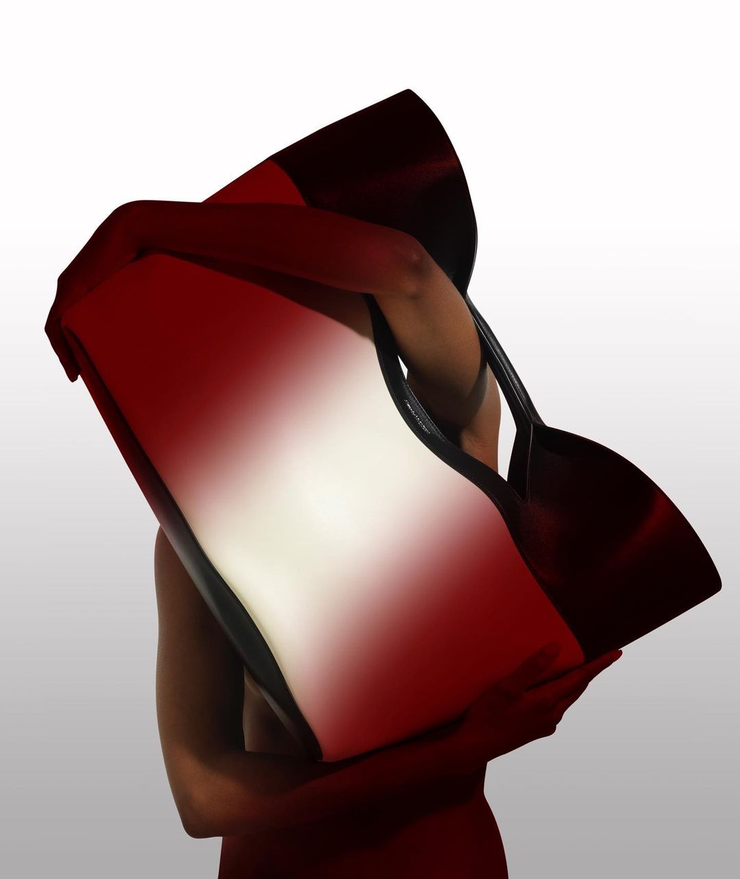 An adult holds a large tote bag with a red-to-white gradient against a white background in a studio shot with soft, even lighting.