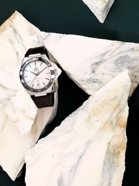 Omega Constellation wristwatch with a black leather strap on white marble fragments against a dark green background in a still life.
